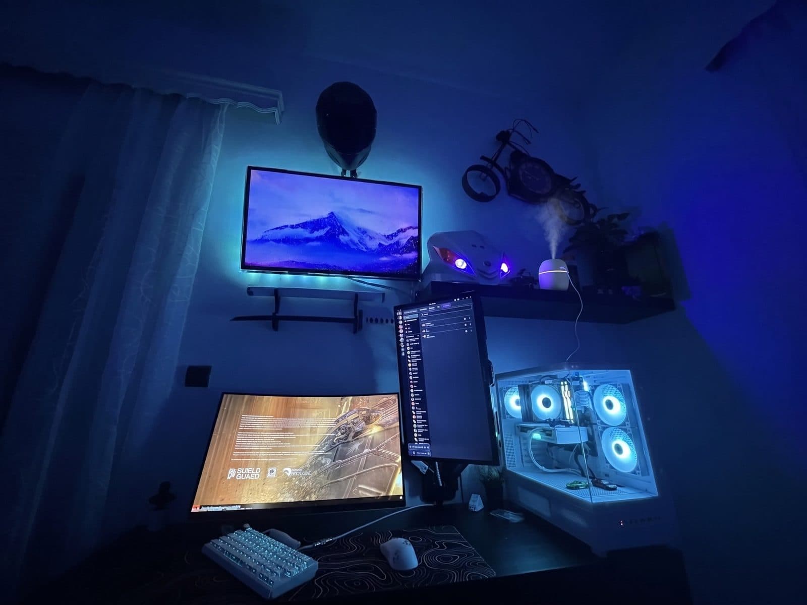 Blue lit gaming room with an LED strip behind a wall mounted TV, a white PC and a humidifier misting on a shelf